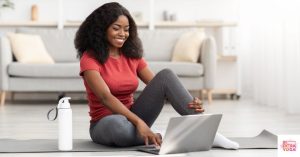 Woman on yoga mat with water bottle and looking at laptop. This article discusses how to find an online wellness coach.