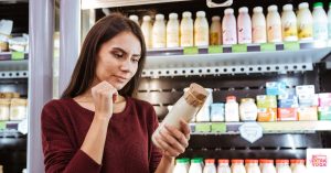 Woman reading food label on milk in front of dairy case at grocery store. This article discusses elimination diet reintroduction.