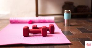 Pink yoga mat on tile floor with free weights and tumbler on the ground. This articles asks: Should I do yoga before or after a workout?