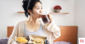 Woman drinking soda and eating a calzone on edge of bed. This article discusses which hormonal imbalance foods to avoid.
