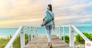 Woman walking to the beach holding a yoga mat. This article discusses the best exercise for gut health.