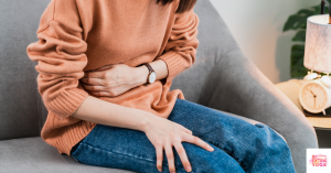 Woman in orange sweater sitting on couch and holding stomach. This article asks can hormone imbalance cause inflammation?