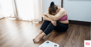 Woman in workout clothes and sitting on floor with head down. This article discusses what losing weight with a hormonal imbalance takes.